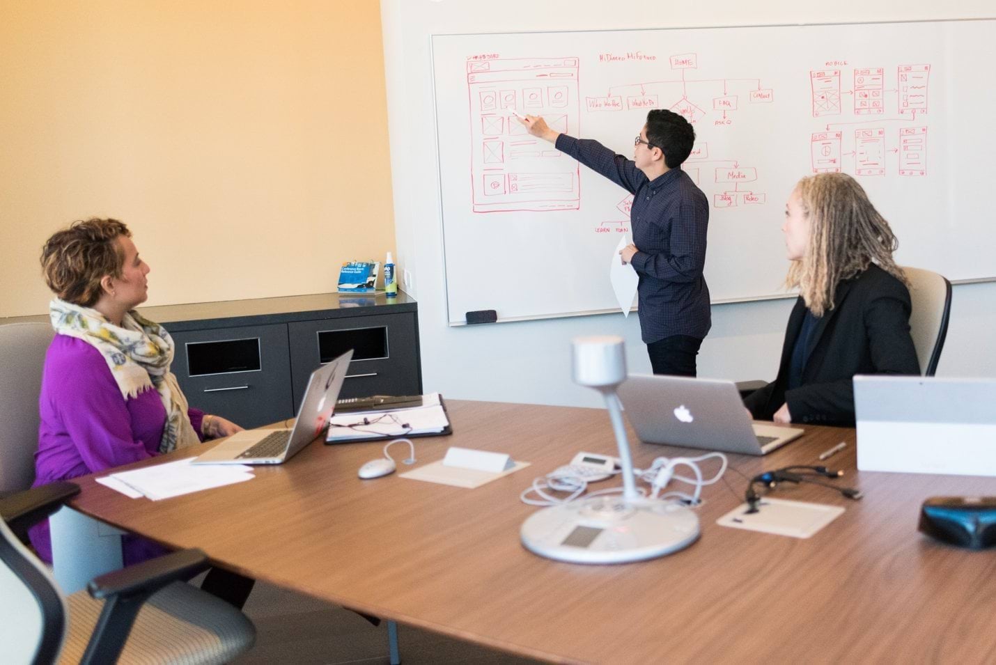 Two women at a table watch a man write on a whiteboard